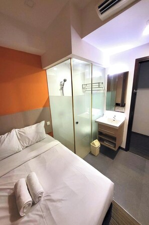 Una habitación de hotel con cama, ducha envuelta en vidrio, lavamanos y espejo.