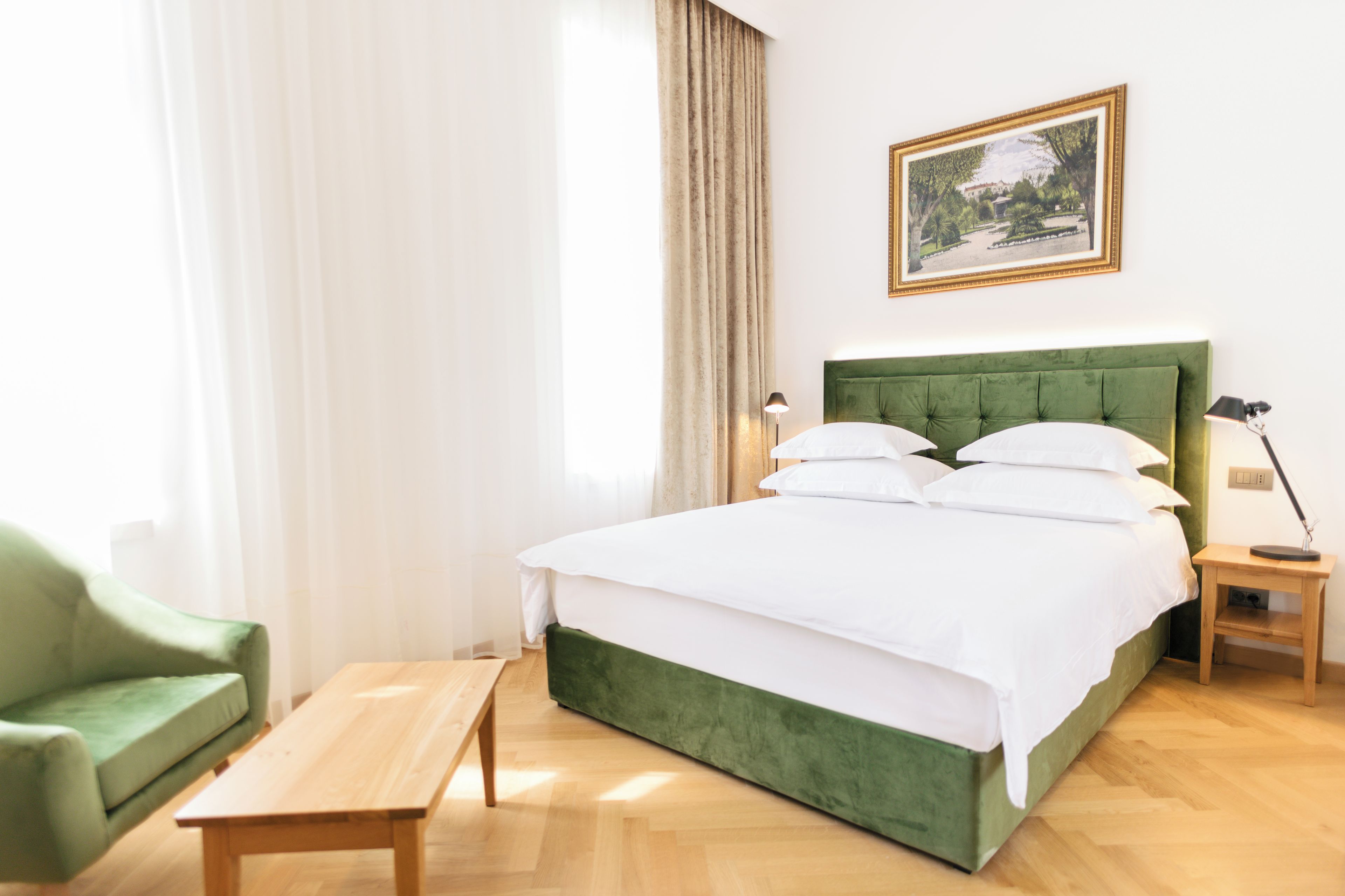 Ein ordentlich eingerichtetes Hotelzimmer mit einem mit grünem Stoff bezogenen Bett, einem Holfnachtschränkchen, einer Nachttischlampe, einem grünen Sessel und einer Holzbank.