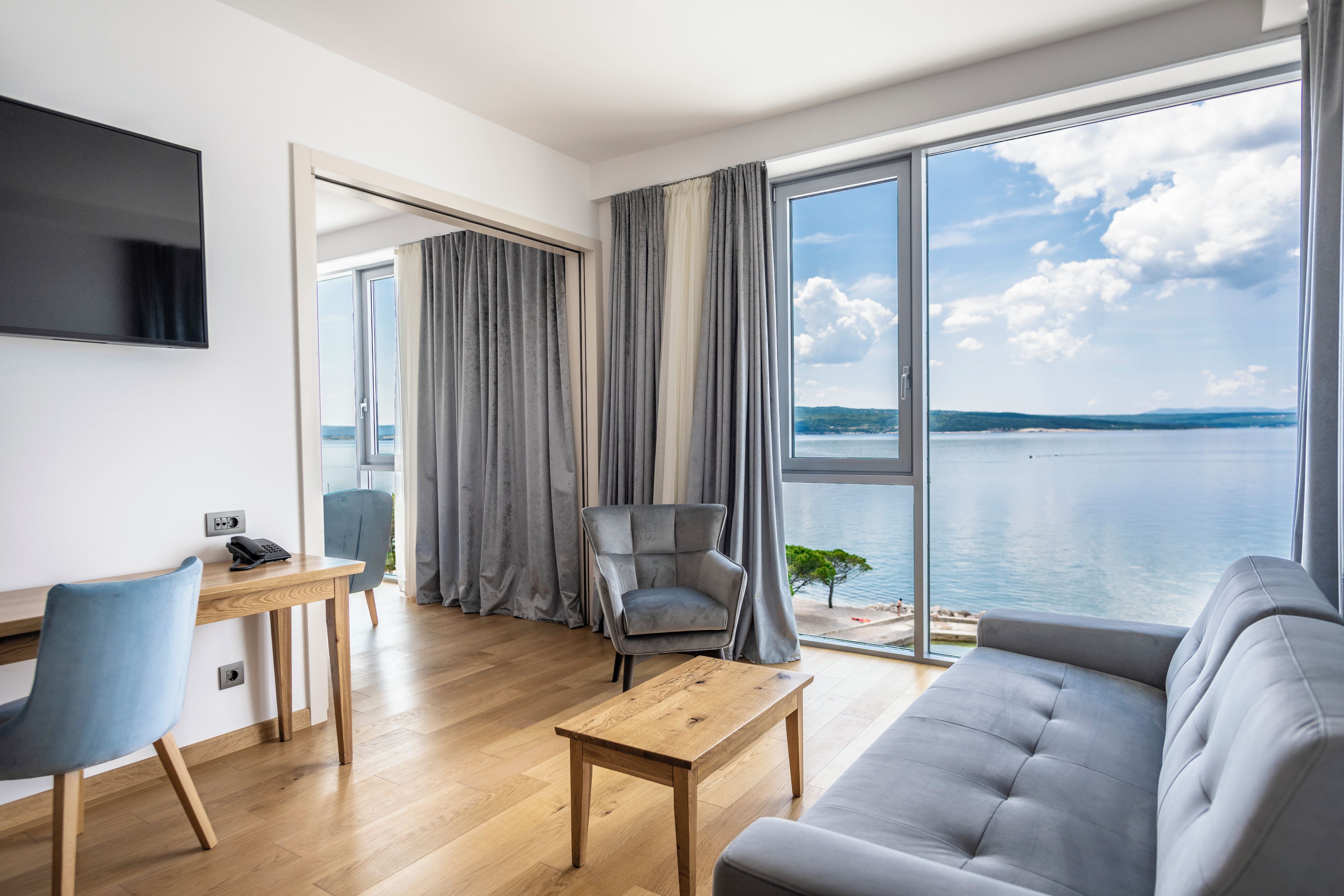 A modern living room with a sofa, a coffee table, a desk with a chair, a television, and a large window with a view of the sea.