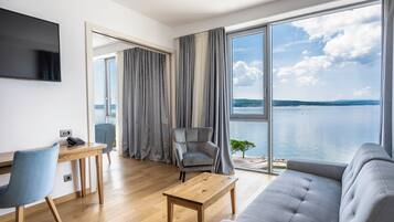 A modern living room with a sofa, a coffee table, a desk with a chair, a television, and a large window with a view of the sea.