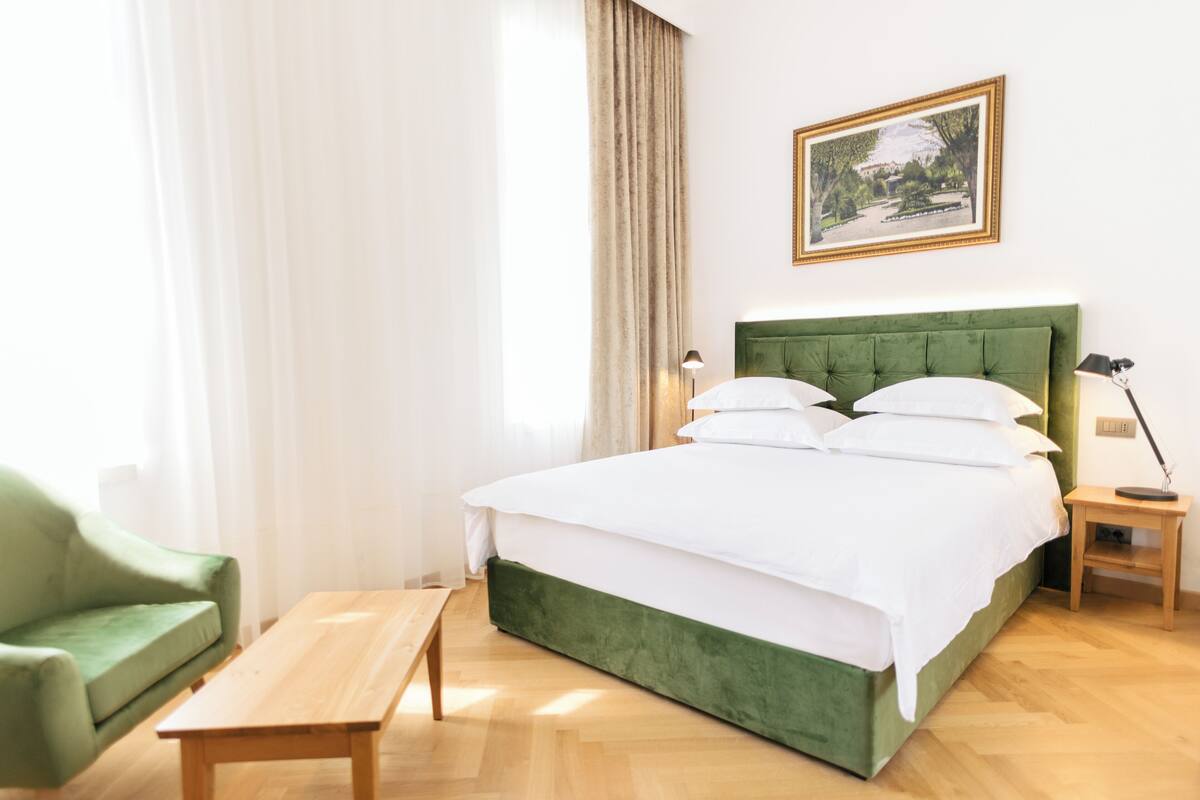 Siististi järjestetty hotellihuone, jossa on vihreäverhoiltu sänky, puinen yöpöytä, sängyn vieressä oleva lamppu, vihreä nojatuoli ja puinen penkki.