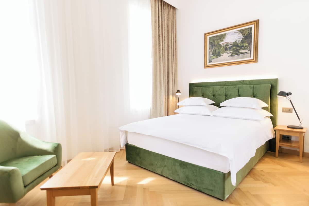 A neatly arranged hotel room with a green upholstered bed, a wooden nightstand, a green armchair, and a wooden table.
