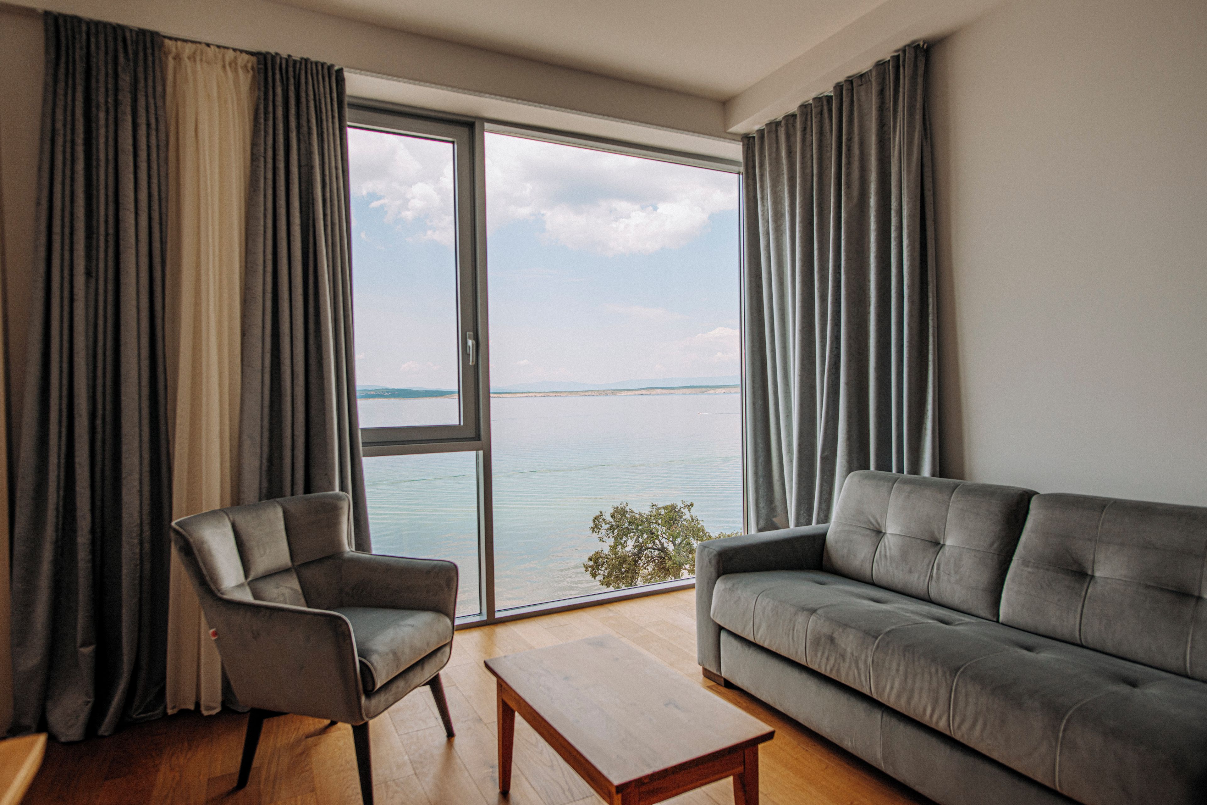 Ein modernes Wohnzimmer mit Sofa, Sesseln und Couchtisch, großen Fenstern mit Vorhängen und Blick aufs Meer.