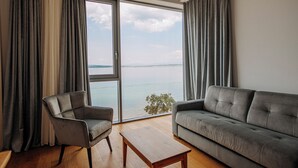 Ένα μοντέρνο σαλόνι με έναν καναπέ, μια πολυθρόνα και ένα τραπεζάκι σαλονιού, μεγάλα παράθυρα με κουρτίνες και θέα στη θάλασσα.