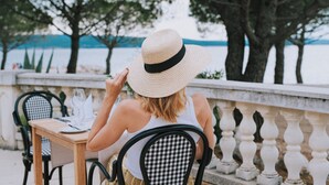 Ένα άτομο κάθεται σε ένα τραπέζι σε εξωτερικό χώρο, με θέα δέντρα και μια έκταση νερού.