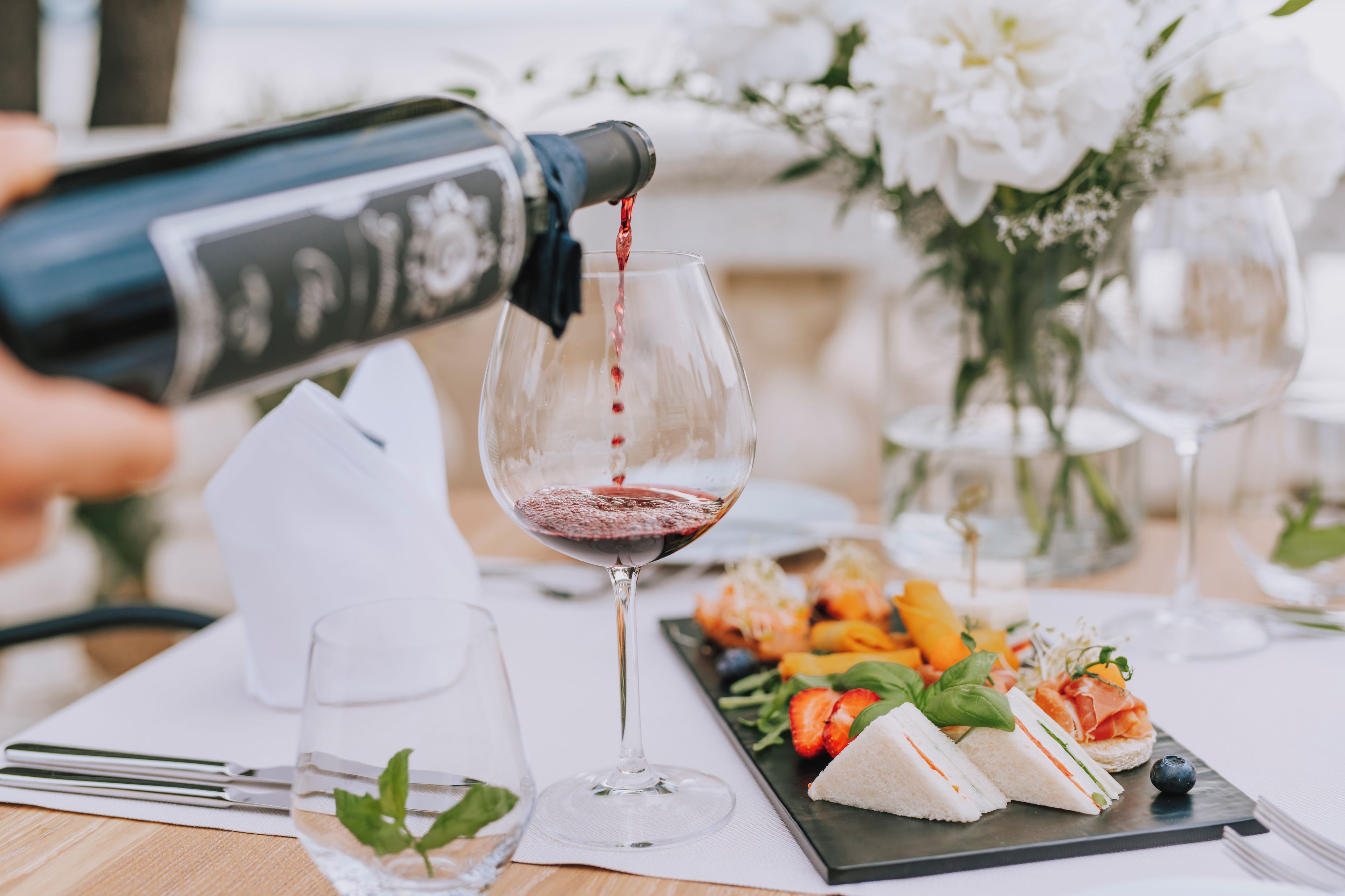 Eine Weinflasche wird in ein Glas eingeschenkt, dazu ein Teller mit Sandwiches und eine Blumenkomposition auf einem Tisch.