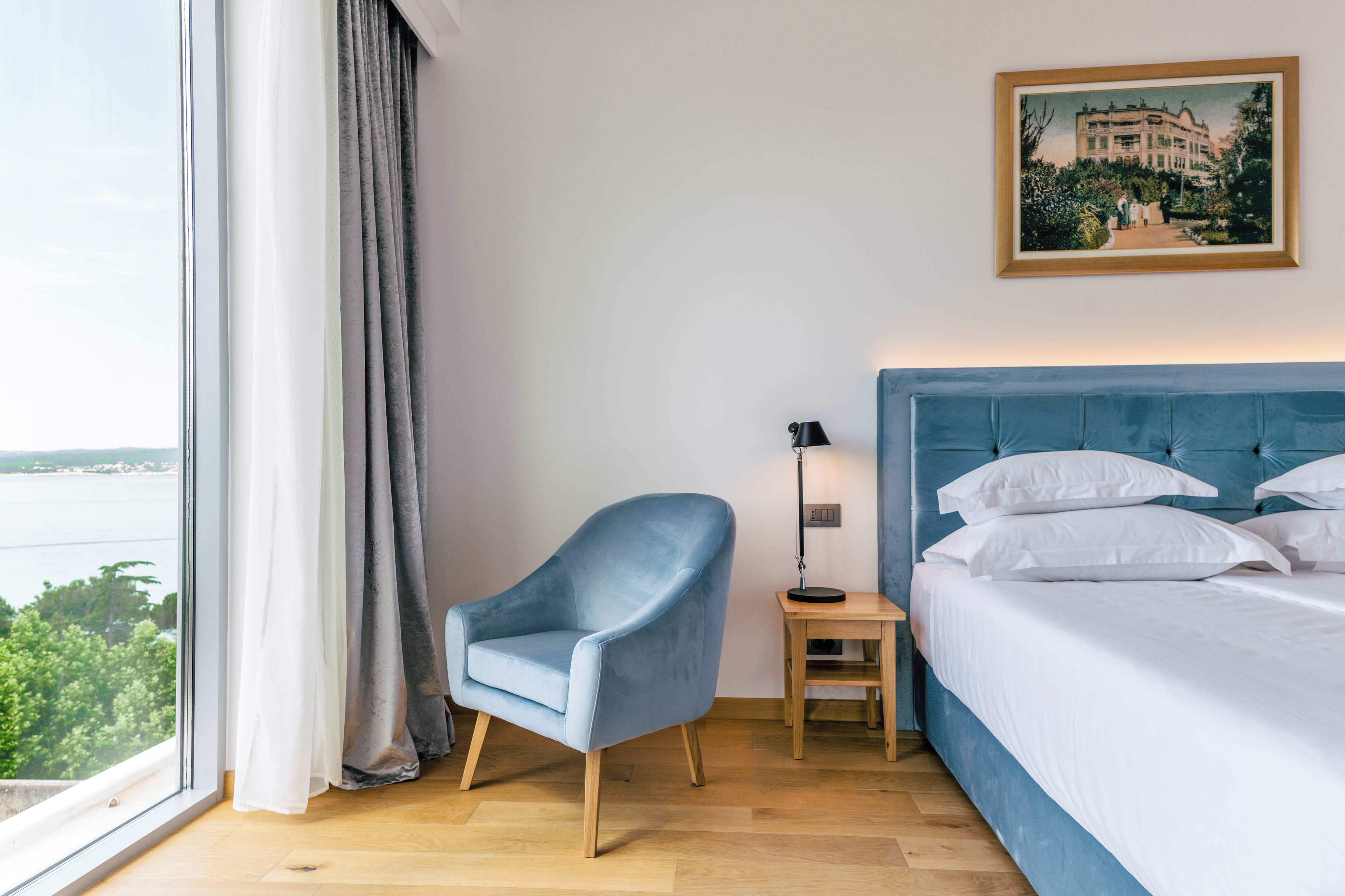 A hotel room with a large window, a blue armchair, a bedside table with a lamp, and a bed with white linens.