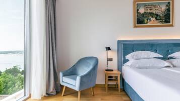 A hotel room with a large window, a blue armchair, a bedside table with a lamp, and a bed with white linens.