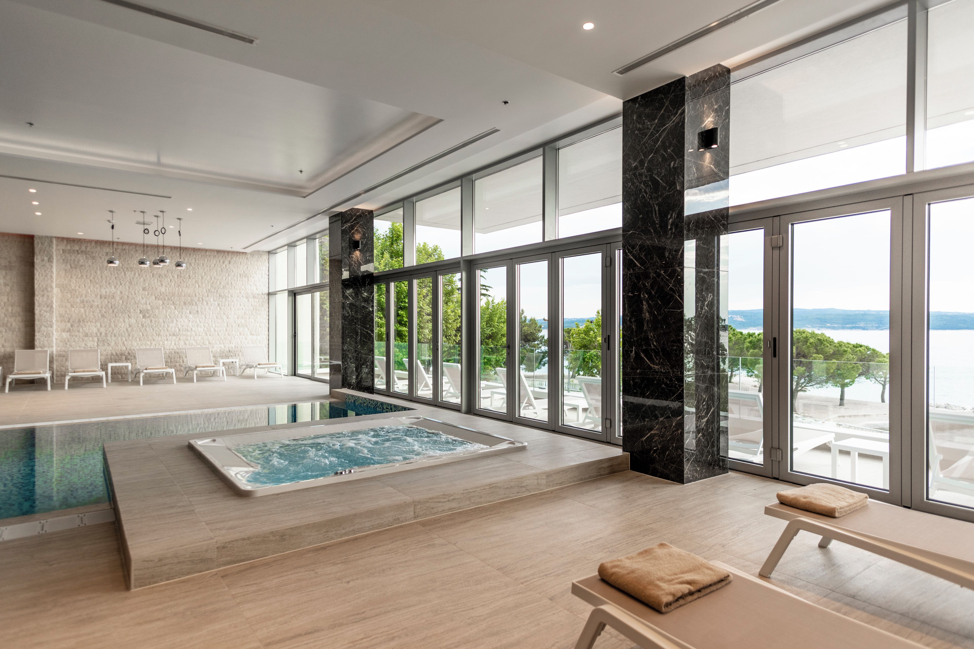 A modern indoor swimming pool with a view of the sea, surrounded by large windows and a seating area with chairs.