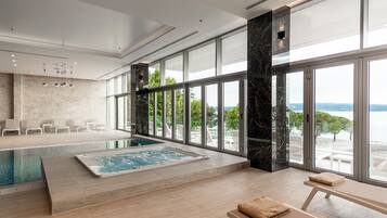 A modern indoor swimming pool with a view of the sea, surrounded by large windows and a seating area with chairs.