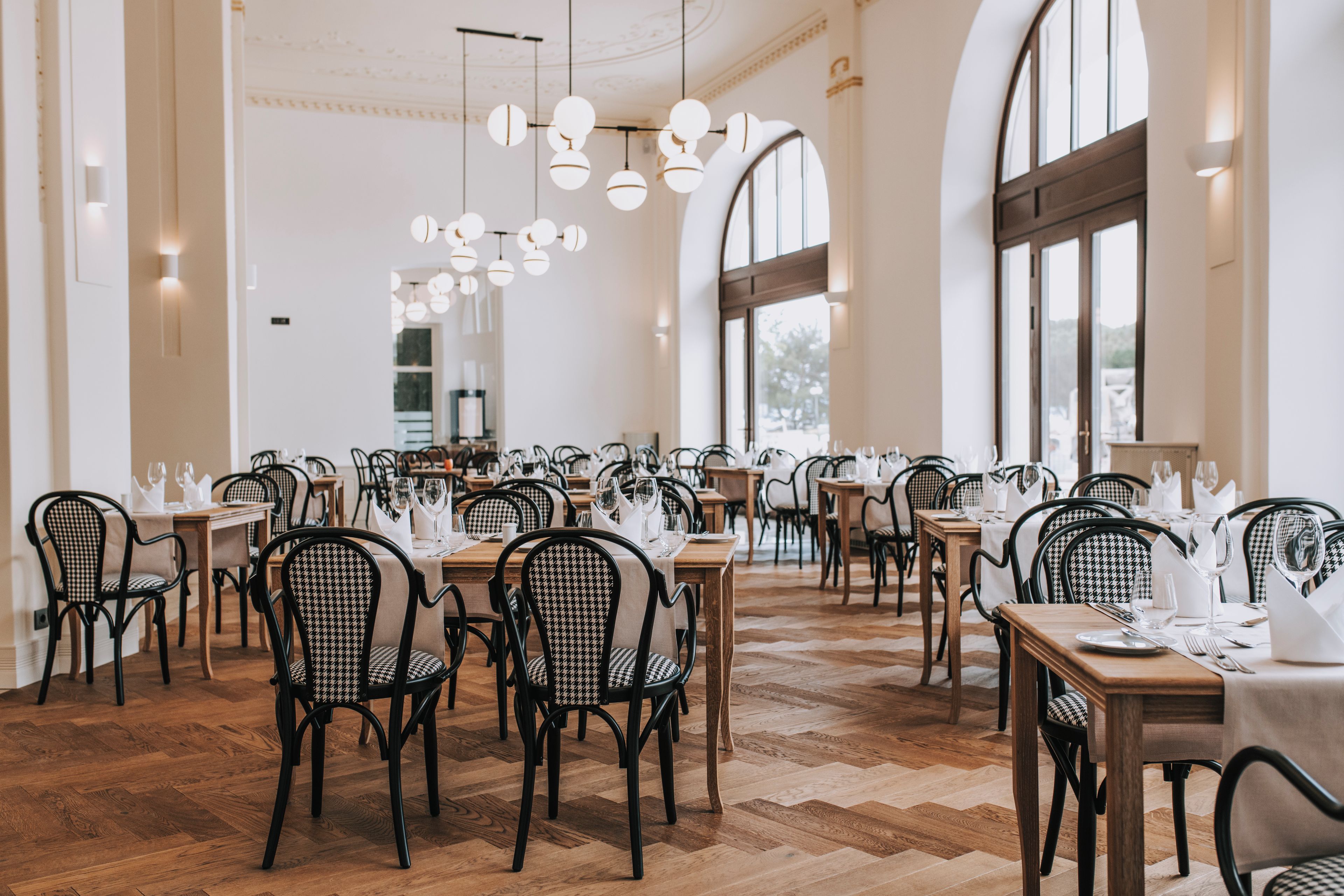 Ein Restaurant mit mehreren Tischen, Stühlen, großen Fenstern und hängenden Lampen.