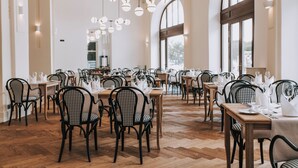 Ένα εστιατόριο με πολλά τραπέζια και καρέκλες, μεγάλα παράθυρα και κρεμαστά φωτιστικά.