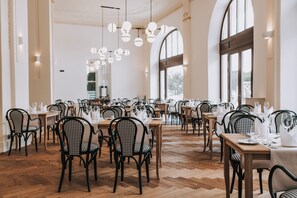 Ένα εστιατόριο με πολλά τραπέζια και καρέκλες, μεγάλα παράθυρα και κρεμαστά φωτιστικά.
