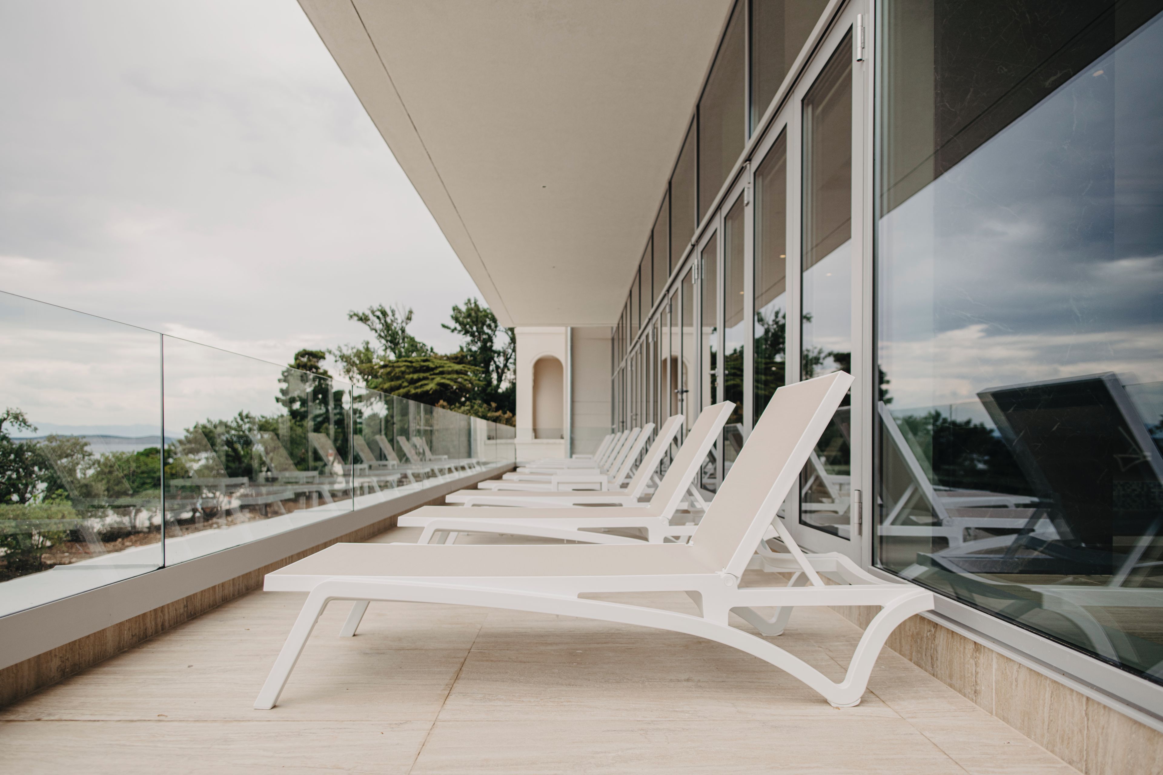 A modern outdoor lounge area with white lounge chairs and a glass balustrade overlooking a scenic landscape.