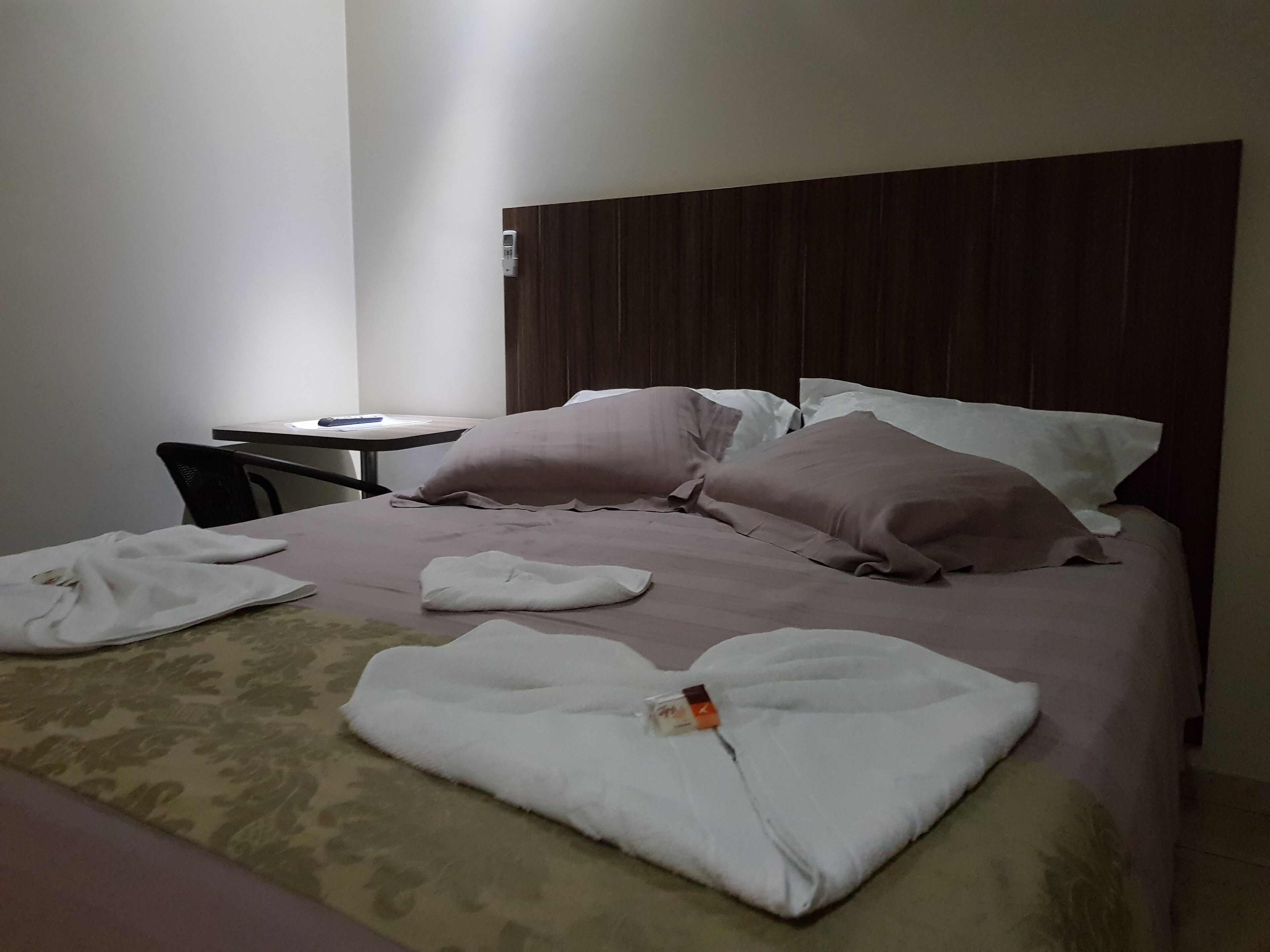 Una cama bien hecha con almohadas y una toalla doblada, una mesita con un control remoto y una silla en una habitación de hotel.