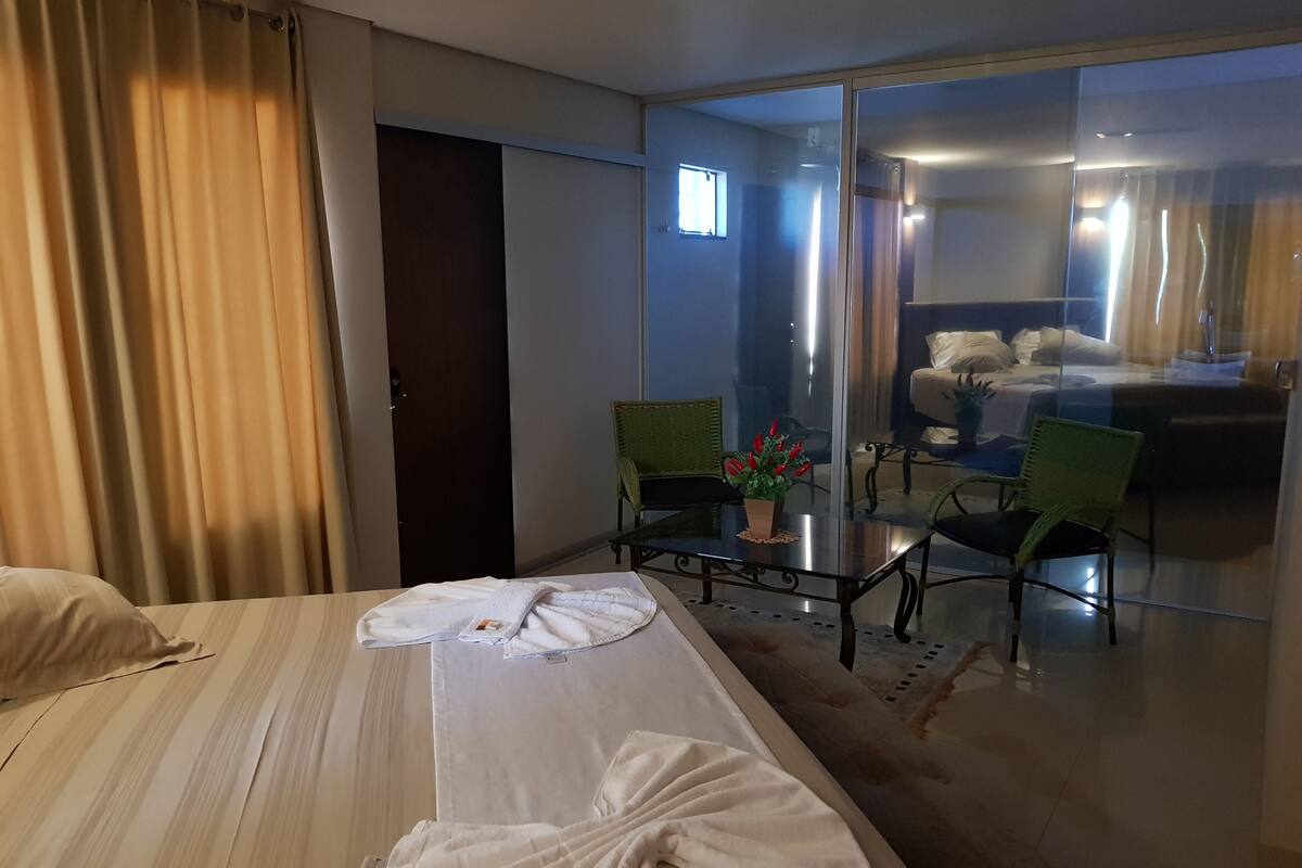 A hotel room with a bed, a glass partition, a small table with chairs, and a view of another room with a bed and a chair.
