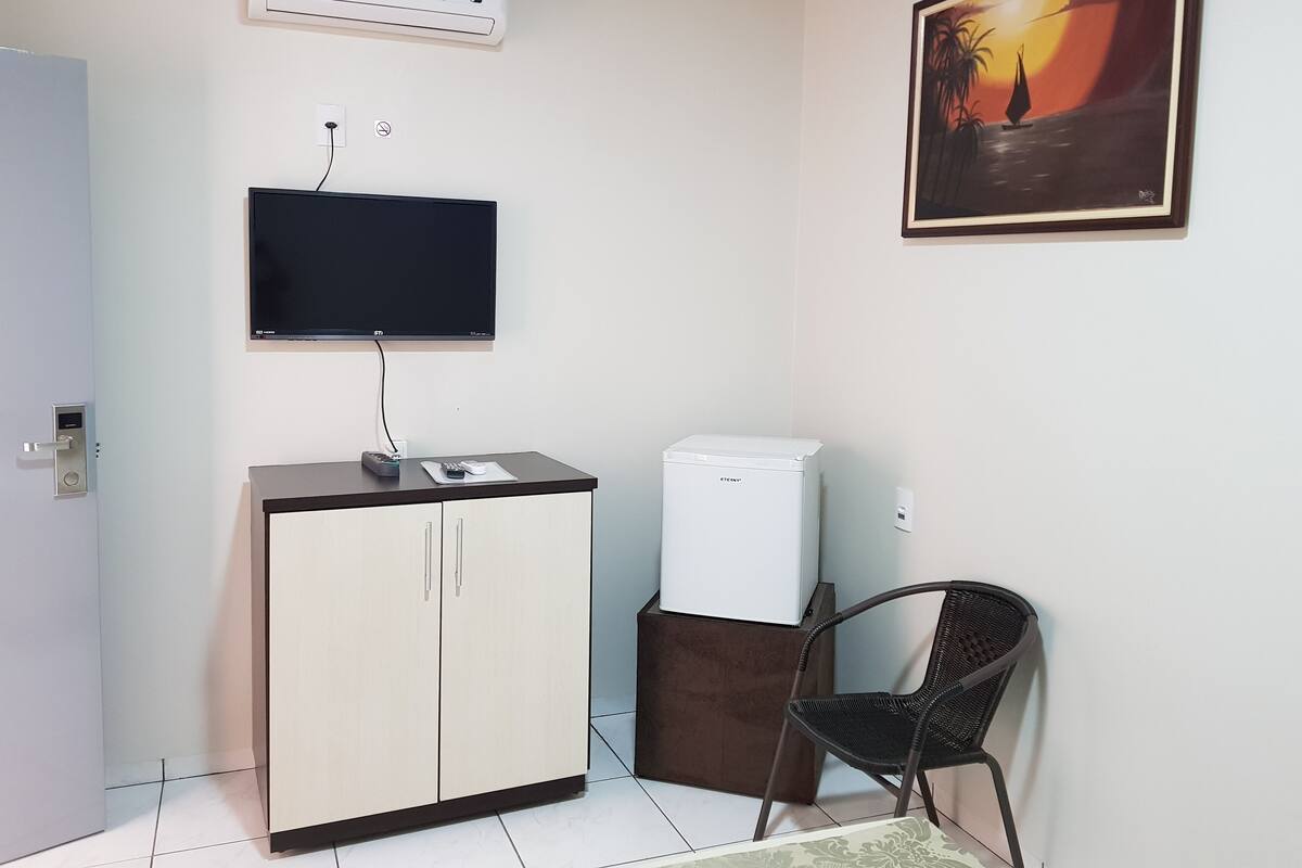 A small room with a TV mounted on the wall, a cabinet with a remote control, a small refrigerator, and a chair.