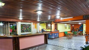 A reception area with a wooden reception desk, tiled floor, and a seating area with chairs and tables. - Chaua Porto Hotel (Porto Seguro)