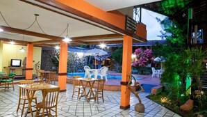 An outdoor dining area with wooden tables and chairs, a pool, and a covered seating area with umbrellas. - Chaua Porto Hotel (Porto Seguro)
