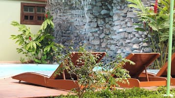 Uma área à beira da piscina com espreguiçadeiras, uma cascata de pedra e vegetação exuberante.