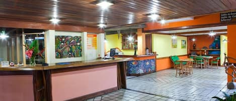 Un área de recepción con un mostrador de madera, suelo de baldosas y un espacio de espera con sillas y mesas.