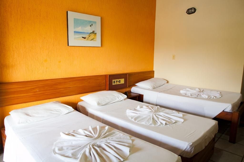 Habitación de hotel con tres camas individuales, cada una con una toalla doblada con cuidado. En la pared, encima de las camas, hay un cuadro de una playa.
