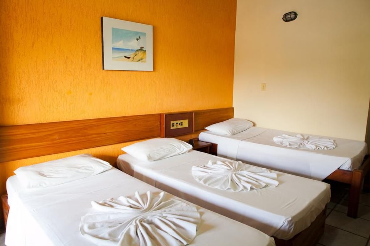 A hotel room with three single beds, each with a neatly folded towel. A painting of a beach scene is on the wall above the beds.