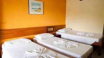 Quarto de hotel com três camas individuais, cada uma com uma toalha dobrada. Uma pintura de uma praia está na parede acima das camas.