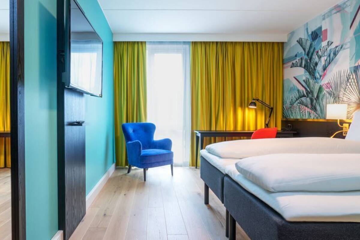 A hotel room with two beds, a blue armchair, and a desk with a lamp.