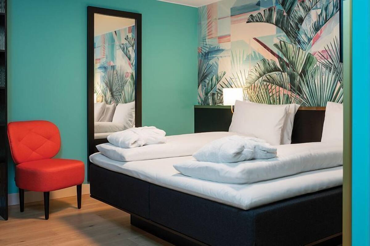 A hotel room with a bed, a red chair, a bookshelf, and a large mirror.