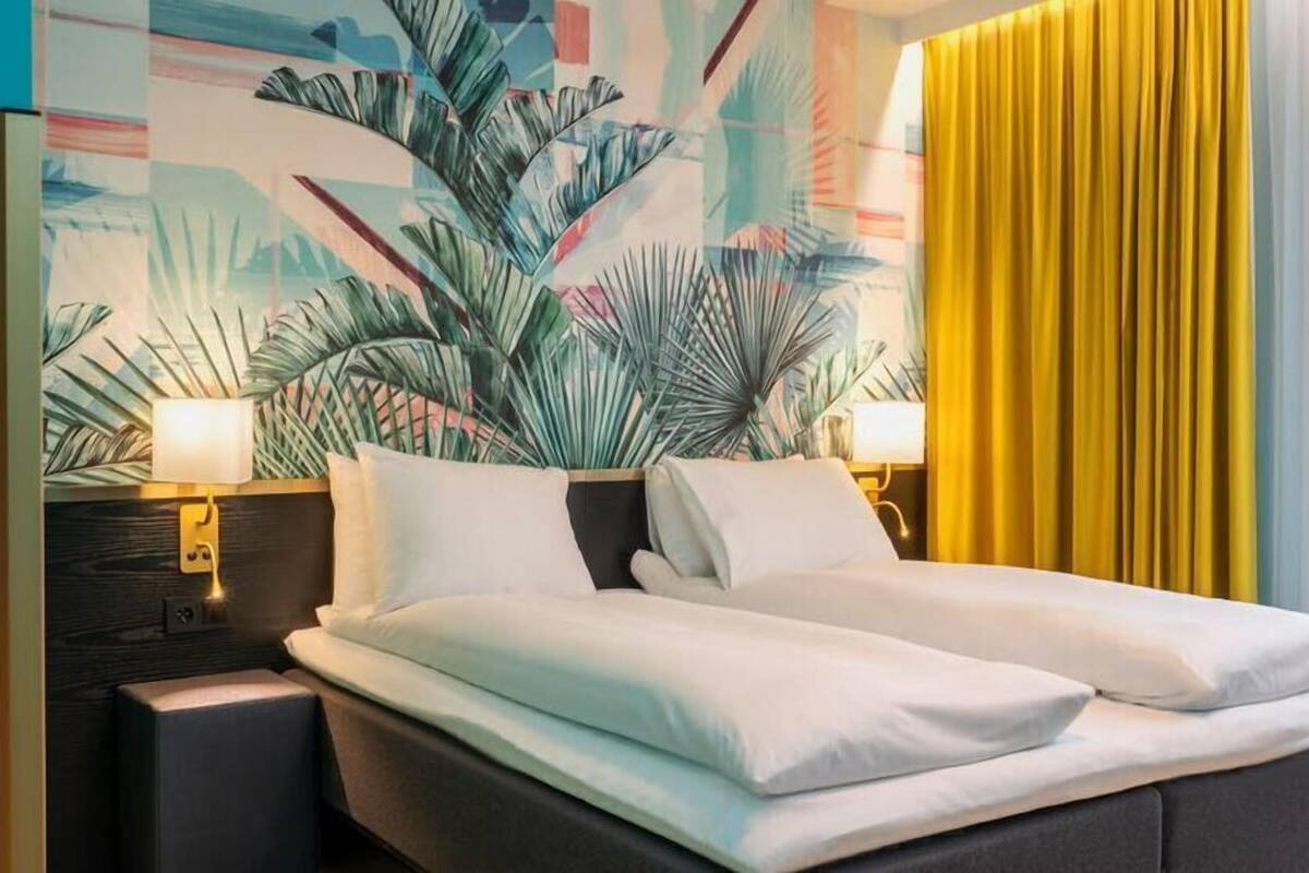 A hotel room with a bed, two bedside lamps, a large tropical-themed wall mural, and a yellow curtain.