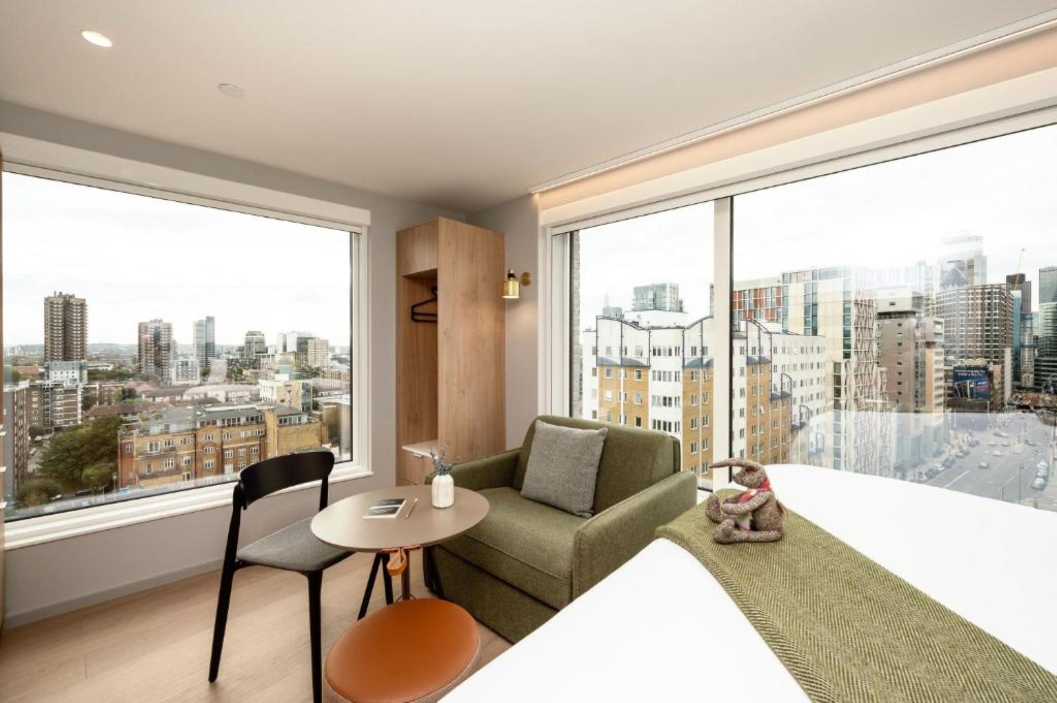 A modern living room with a green sofa, a small table, and a chair. There is a large window offering a city view.