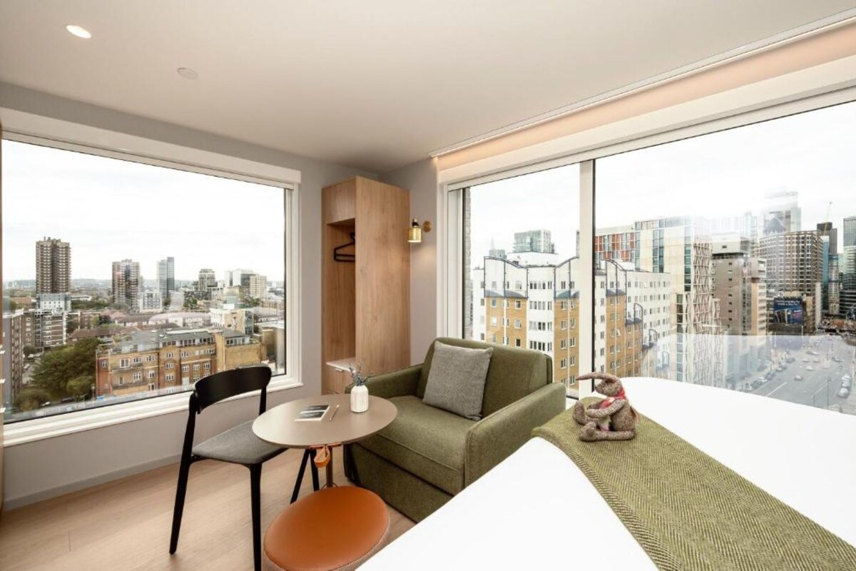 Un salon moderne avec un canapé vert, une petite table et un fauteuil. Une grande fenêtre offre une vue sur la ville.