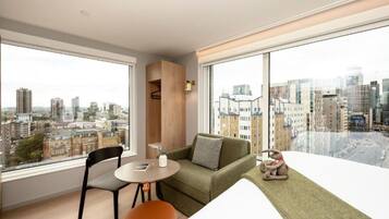 A modern living room with a green sofa, a small table, and a chair. There is a large window offering a city view.