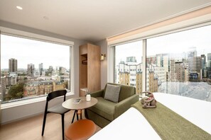 A modern living room with a green sofa, a small table, and a chair. There is a large window offering a city view. - Wilde Aparthotels London Aldgate Tower Bridge (London)
