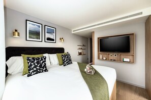 A modern hotel room with a large bed, a flat-screen TV mounted on the wall, and a bedside table with a lamp.
