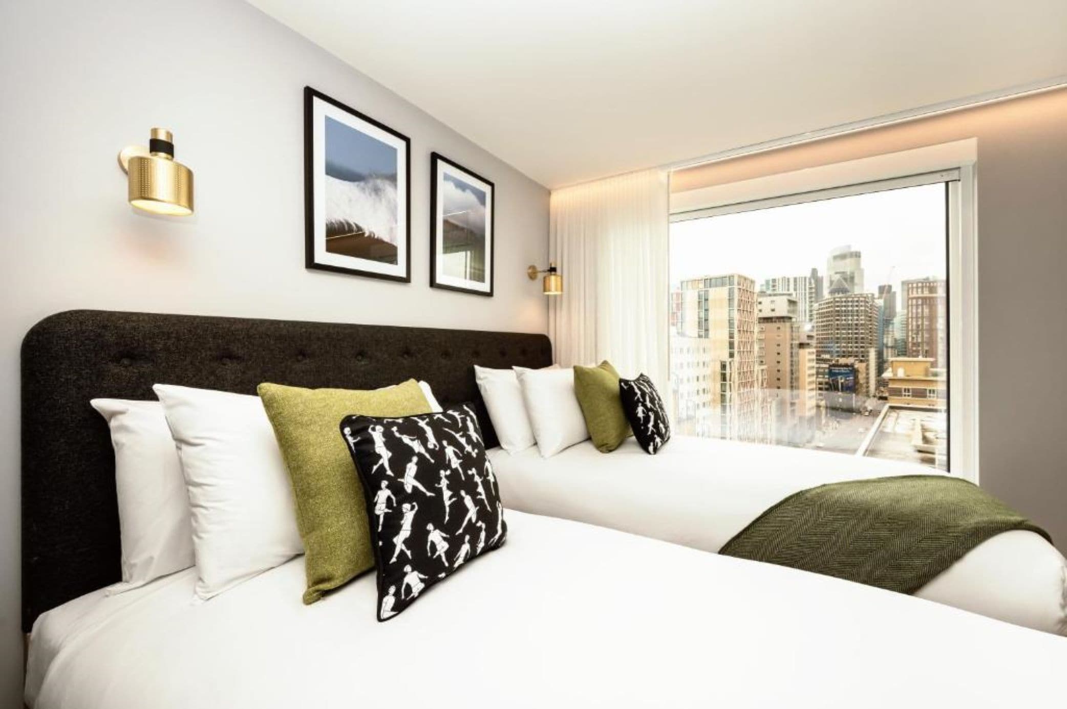 Quarto de hotel com uma cama grande, duas fotos emolduradas na parede e vista da cidade através de uma janela ampla.