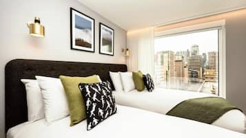 Quarto de hotel com uma cama grande, duas fotos emolduradas na parede e vista da cidade através de uma janela ampla.