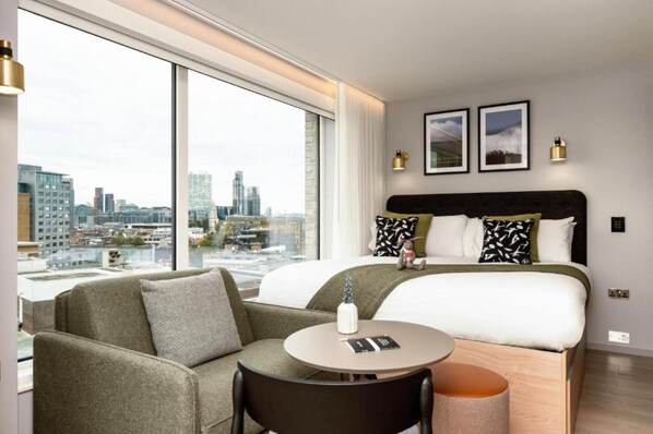 A modern hotel room with a large bed, a sofa, a round table, and a city view. - Wilde Aparthotels London Aldgate Tower Bridge (London)