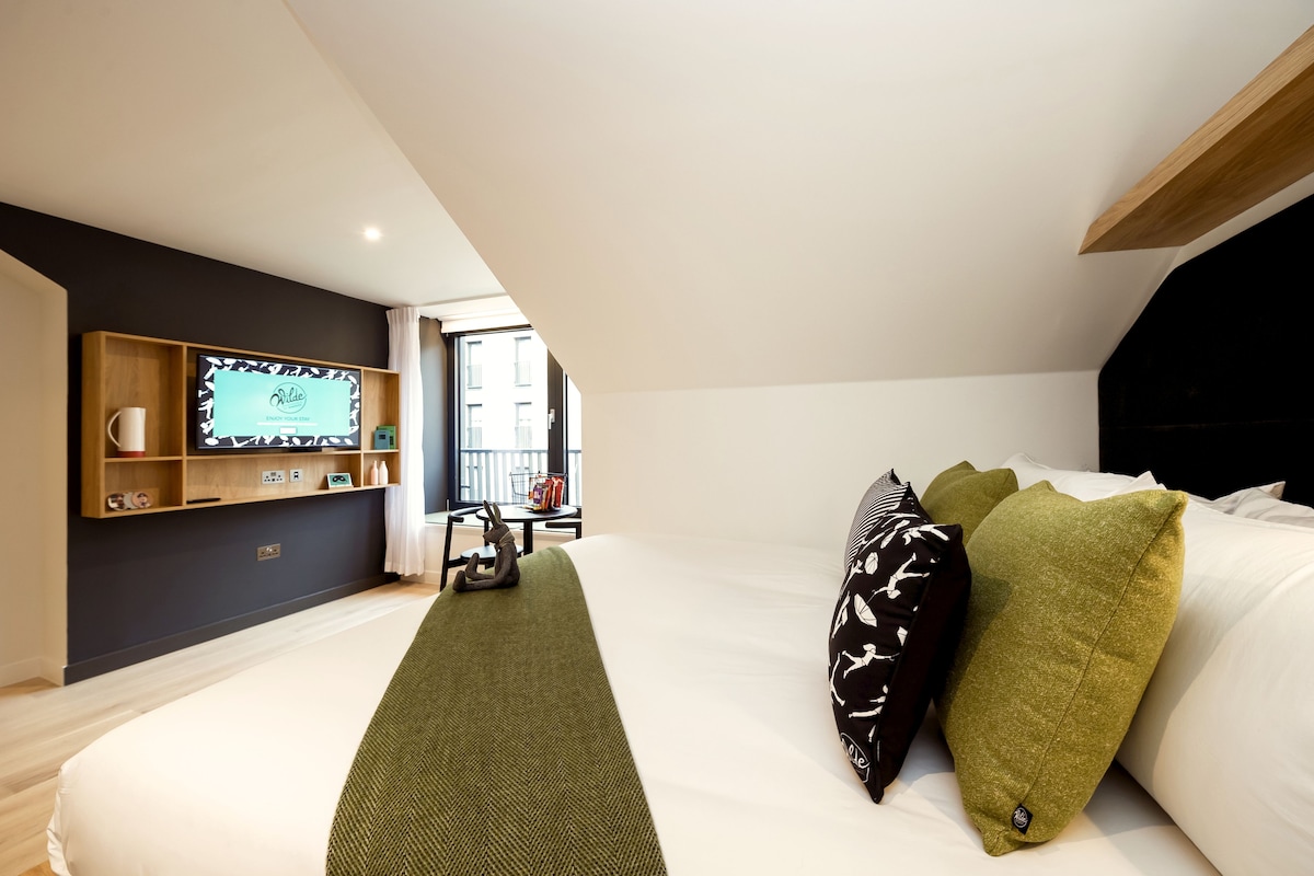 A modern bedroom with a large bed, a wooden shelf with a television, and a window providing a view of the outside.