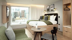 Et moderne hotelværelse med en stor seng, et lille rundt bord, en sofa og udsigt over bybilledet.
