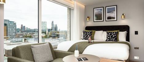 Quarto de hotel moderno com uma cama grande, um sofá, uma mesa redonda e vista para a cidade através de amplas janelas.