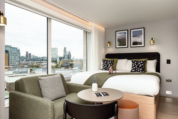 A modern hotel room with a large bed, a sofa, a round table, and a view of the cityscape through large windows. - Wilde Aparthotels London Aldgate Tower Bridge (London)