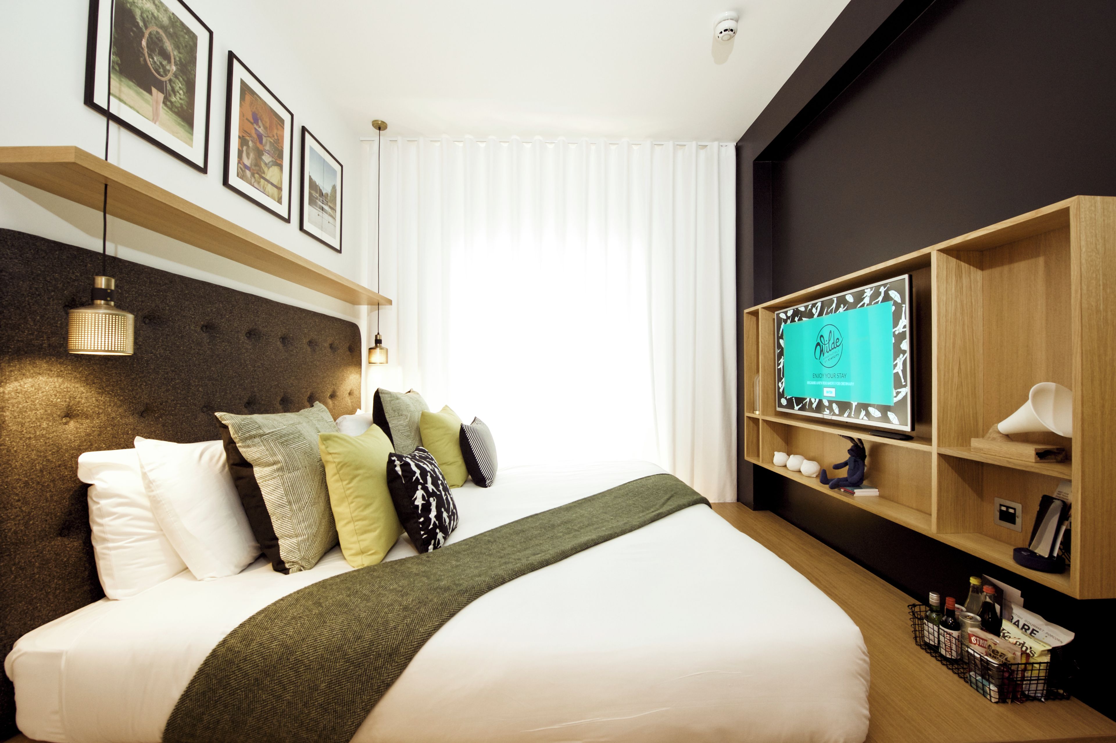 A hotel room with a large bed, a mounted TV, and a shelf with various items.