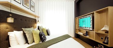 A hotel room with a large bed, a mounted TV, and a shelf with various items.