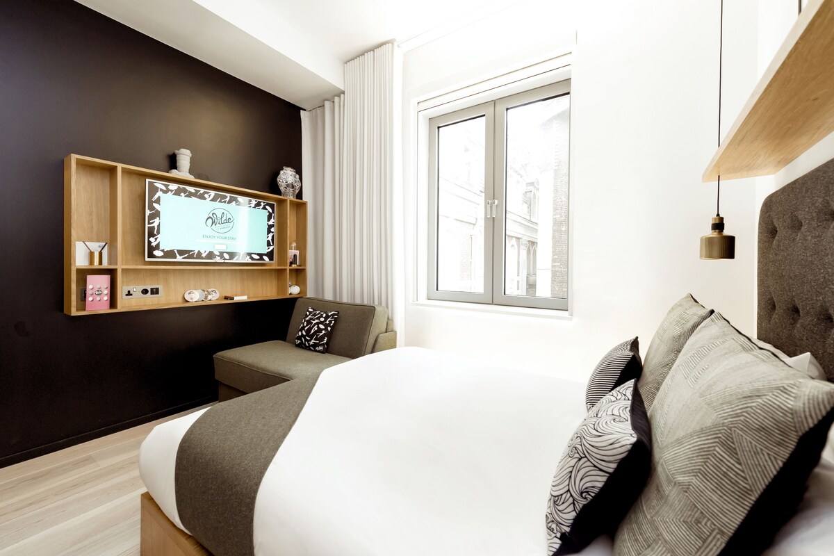 A modern hotel room with a large bed, a TV mounted on the wall, a sofa, and a window with curtains.