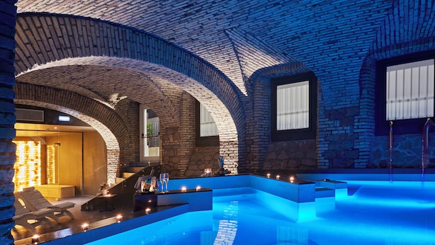 A modern indoor swimming pool with a brick arch ceiling, a lounge area with a sofa and table, and a stone wall.
