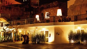 Pátio de hotel à noite, com muro de pedra, plantas em vasos e uma varanda iluminada.