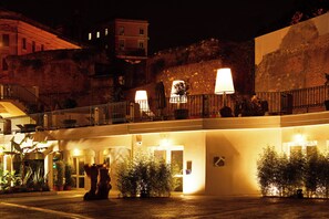 A hotel courtyard at night with a stone wall, potted plants, and a balcony with lights. - The Building (Rome)