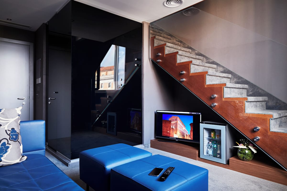 A modern living room with a glass door leading to a balcony, a staircase with a wooden finish, a flat-screen TV, and a mini-fridge.